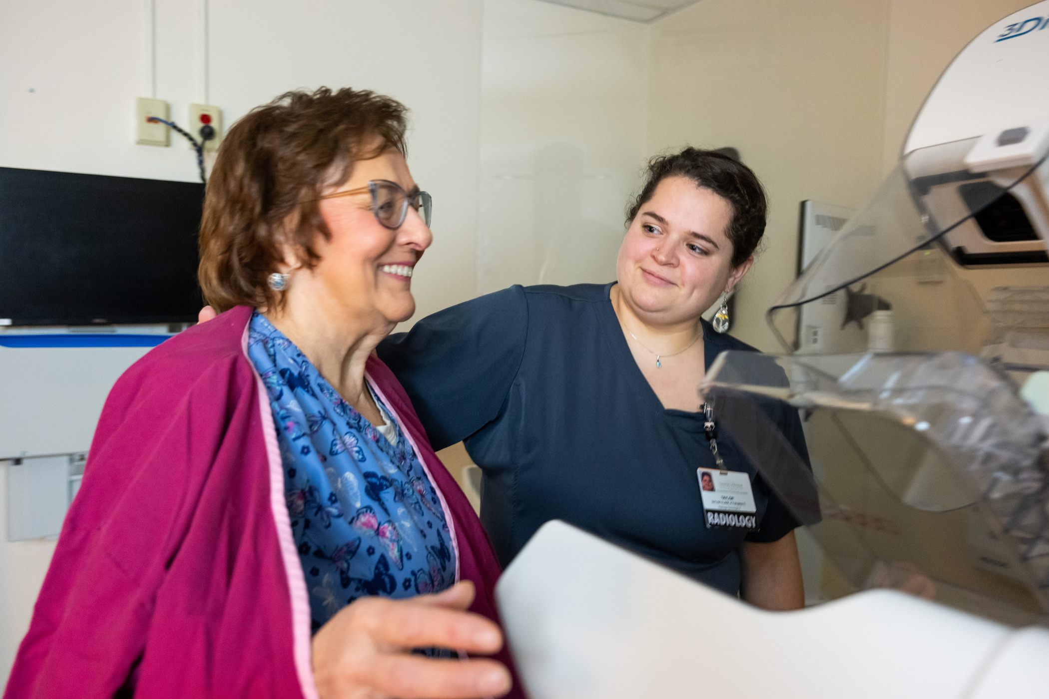 Radiology staff member assisting a patient next to a mammography machine in an imaging exam room