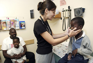 Pediatrician holding young boy's face, as to provide tonsil exam. Young boy's father looks on, while sitting, holding his other son.