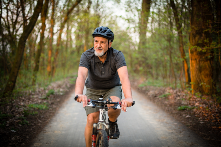 An older man rides his bike on a forested road.