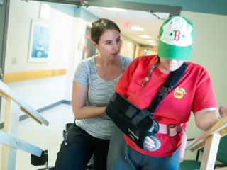 A physical therapist supports a patient wearing arm cast while walking.