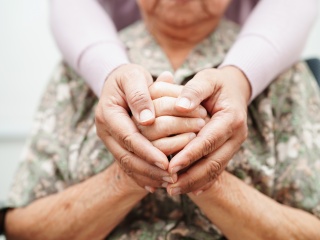 An elderly woman sits while another person wraps their hands around her.