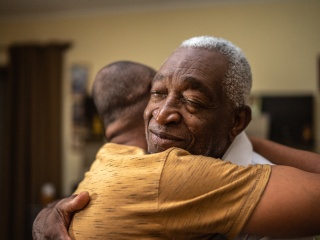 Adult man with grey hair hugging another person.
