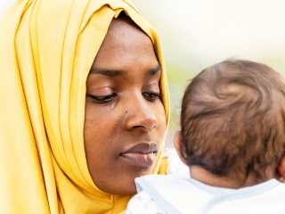 A Black adult woman wearing a yellow head scarf holds an infant.