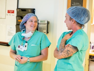 Two nursing staff members in scrubs and hair caps smiling and talking in a hospital operating room hallway.