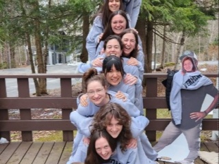 Nine women in matching gray hoodies form a human pyramid on a deck, smiling, next to a cardboard cutout of a person.