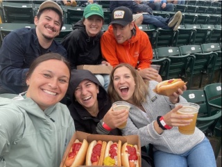 Six OBGYN residents pose for photo in baseball stadium holding hot dogs.