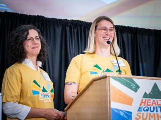 Two smiling event organizers in yellow Health Equity Summit T-shirts stand at a podium bearing the Health Equity Summit logo.