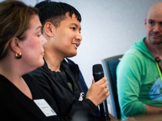 A panelist speaks into a handheld microphone while seated between two other attendees who listen.
