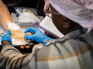 Over-the-shoulder view of a participant wearing blue gloves practicing wound packing and bandaging with gauze and tape on a training limb, with an open notebook nearby.