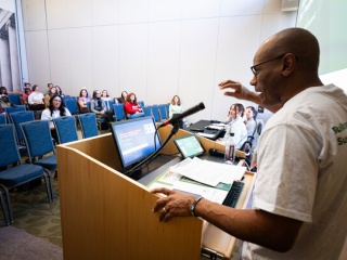A presenter in a white event T-shirt speaks at a podium, gesturing toward an audience seated in an auditorium, with a University of Vermont Health slide projected behind him.