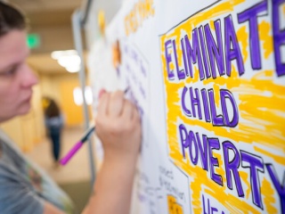 A woman writes on a large poster board with a pink marker. Bold purple and yellow lettering on the board reads “Eliminate Child Poverty.”