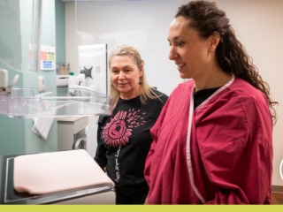 Patient and healthcare professional standing beside a mammography machine in a breast imaging exam room.