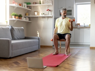 An older man lifts weights while sitting in a chair in his home.