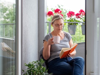 A woman reads a book on a porch with a mug in her hand.