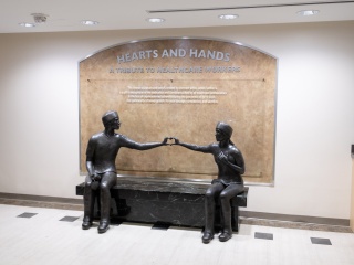 A bronze sculpture of a male and a female provider sitting on a black marble bench making a heart with their hands.