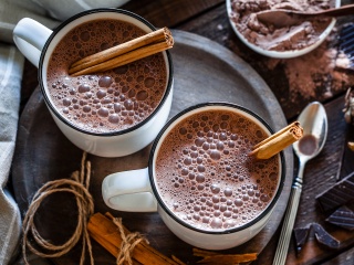 Two mugs of hot chocolate with cinnamon sticks sit on a table.