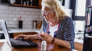 Older woman holding a prescription bottle while looking at her laptop screen.