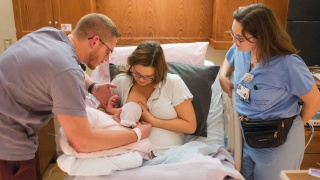 A nurse assists two parents in breastfeeding their newborn baby.