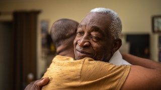 Adult man with grey hair hugging another person.