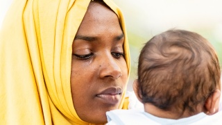 A Black adult woman wearing a yellow head scarf holds an infant.