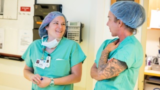 Two nursing staff members in scrubs and hair caps smiling and talking in a hospital operating room hallway.