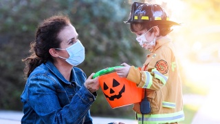 A masked woman gives Halloween candy to child wearing a firefighter costume and a mask.