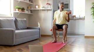An older man lifts weights while sitting in a chair in his home.