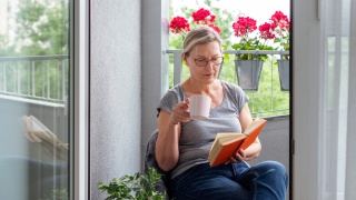 A woman reads a book on a porch with a mug in her hand.