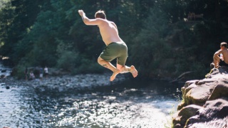 Man jumps off rocks into swimming hole in river.