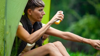 A woman sits outside her tent spraying bug spray on her arm.