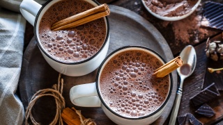 Two mugs of hot chocolate with cinnamon sticks sit on a table.