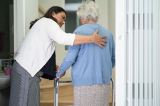 Caregiver helps elderly old woman walk with walker support up the stairs in home.