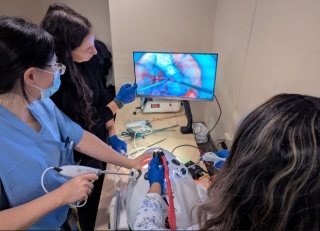 Three OBGYN residents practice a laparoscopic procedure on a training model, with the internal camera view shown on a monitor in front of them