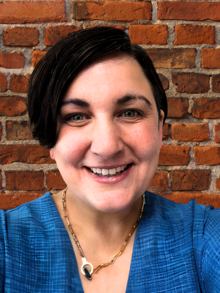 Photo of Christa Hagan-Howe smiling in a blue shirt in front of a brick wall